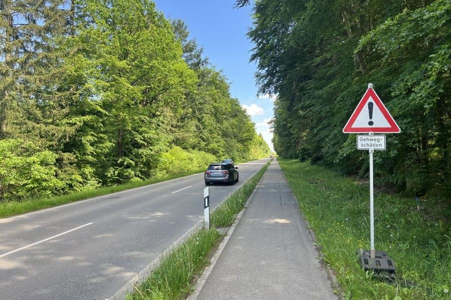 Ein Auto fährt die Flachter Straße entlang, rechts und links sind Büsche und Bäume sowie ein Straßenschild zu sehen.