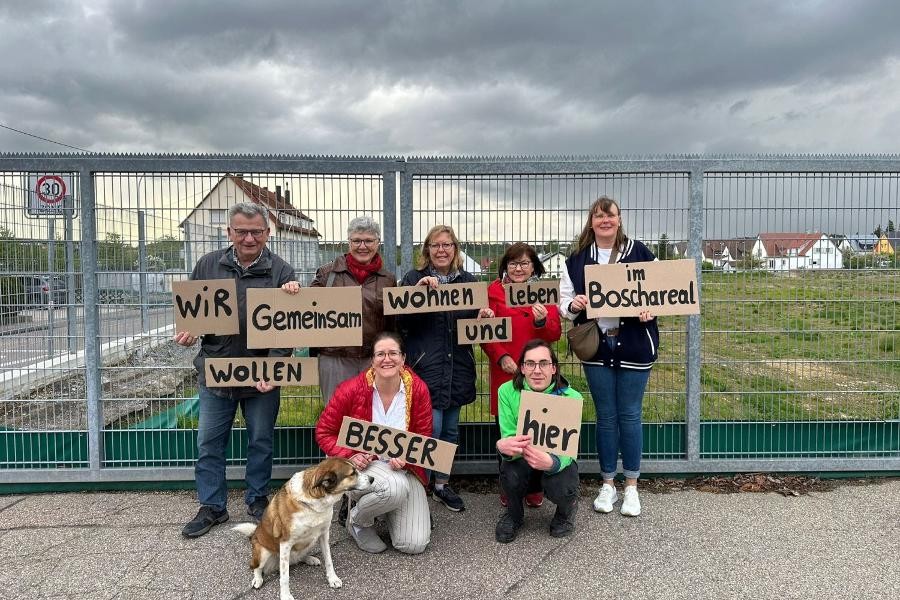 Gruppenbild mit mehreren Personen, die am Zaun des Bosch-Areals stehen und braune Pappschilder mit schwarzen Aufschriften in Händen halten.